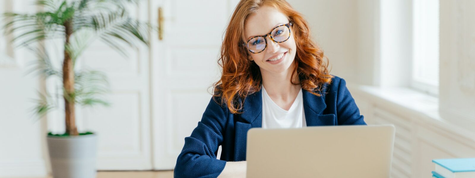 Businesswoman wears glasses for vision correction, sits at desktop in front of opened laptop