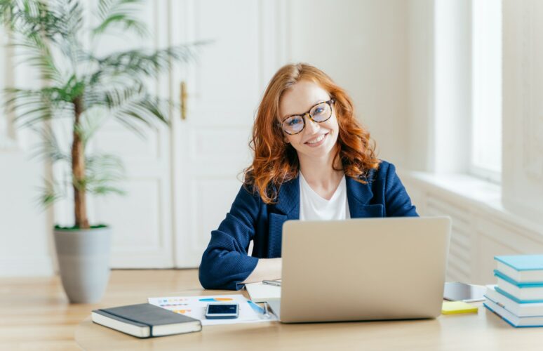 Businesswoman wears glasses for vision correction, sits at desktop in front of opened laptop
